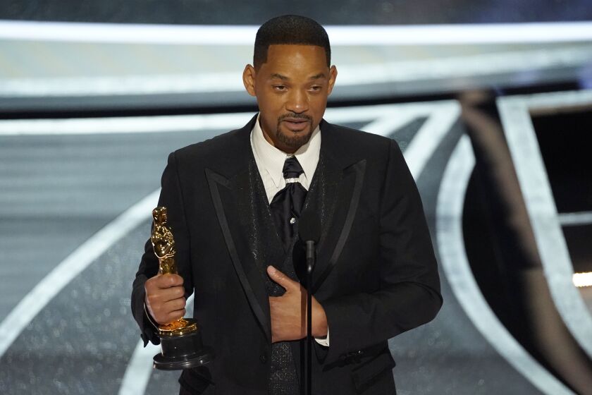 A tearful man with short black hair wearing a black suit, holding a gold Oscar trophy and speaking into a mic on a stage