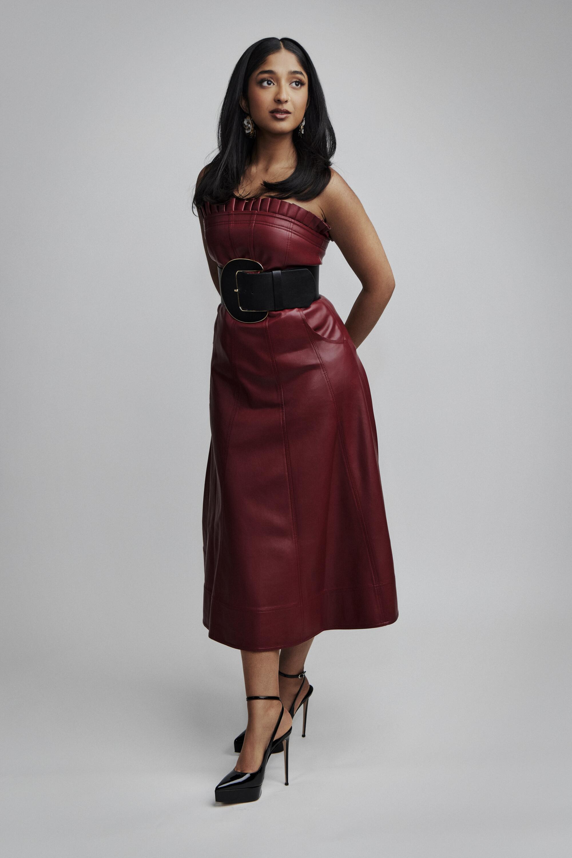 Maitreyi Ramakrishnan stands with her arms behind her back in a strapless maroon dress and heels.