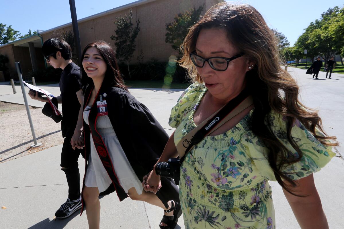 A woman in a graduation gown walks with a young man and a woman on a college campus.