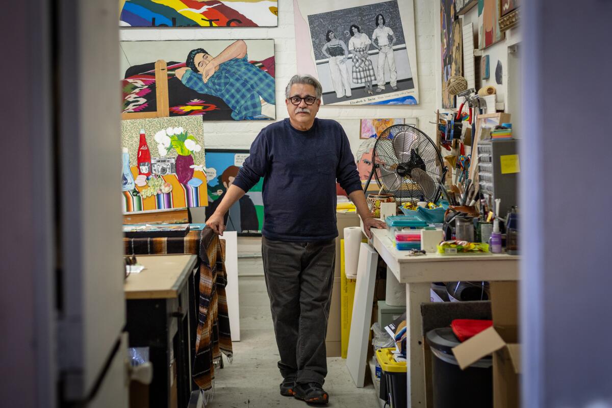 Joey Terrill, a Chicano man in his 60s with a mustache, stands between two tables in a cluttered art studio.