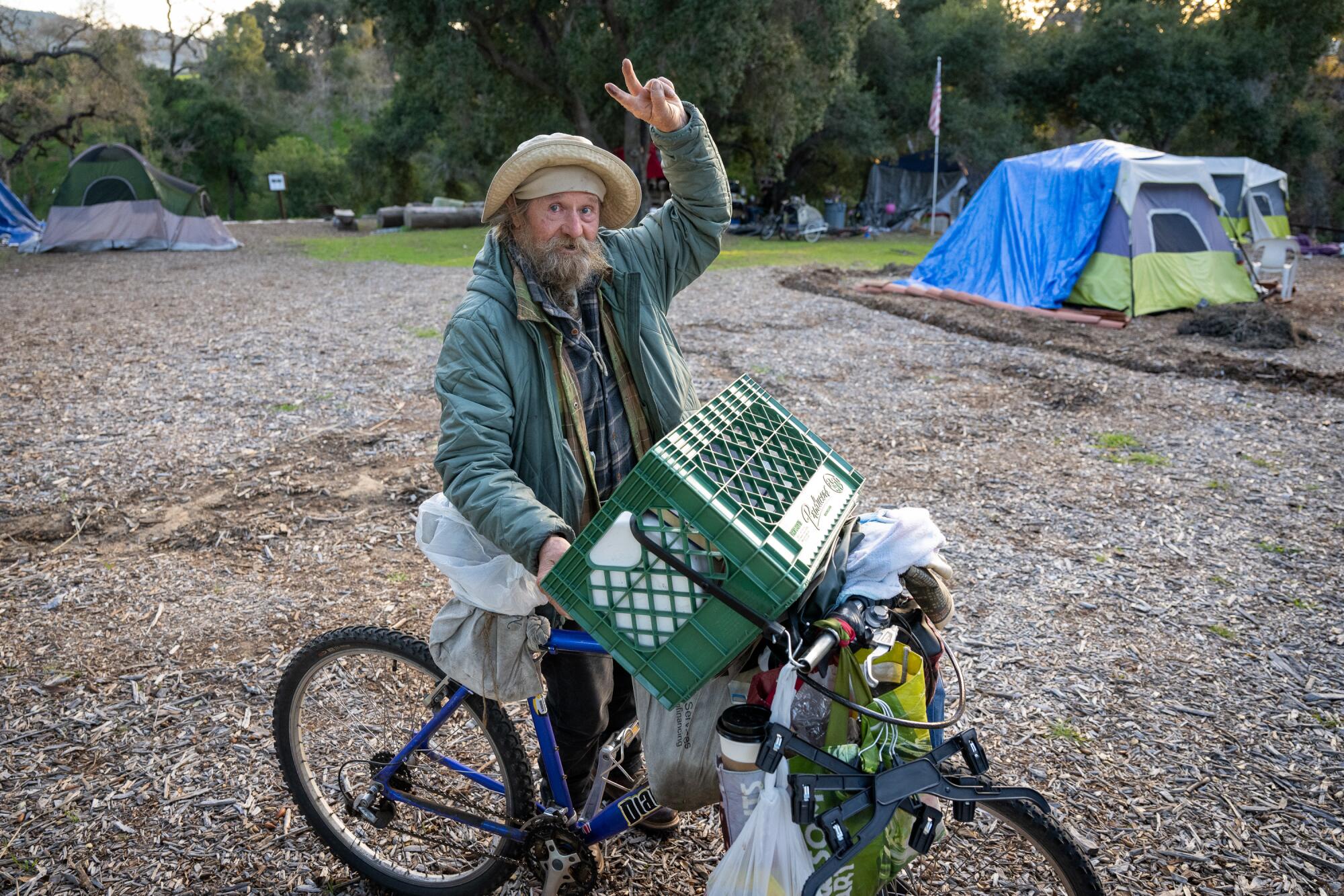 A homeless man on a bike loaded with plastic bags gives the peace sign.