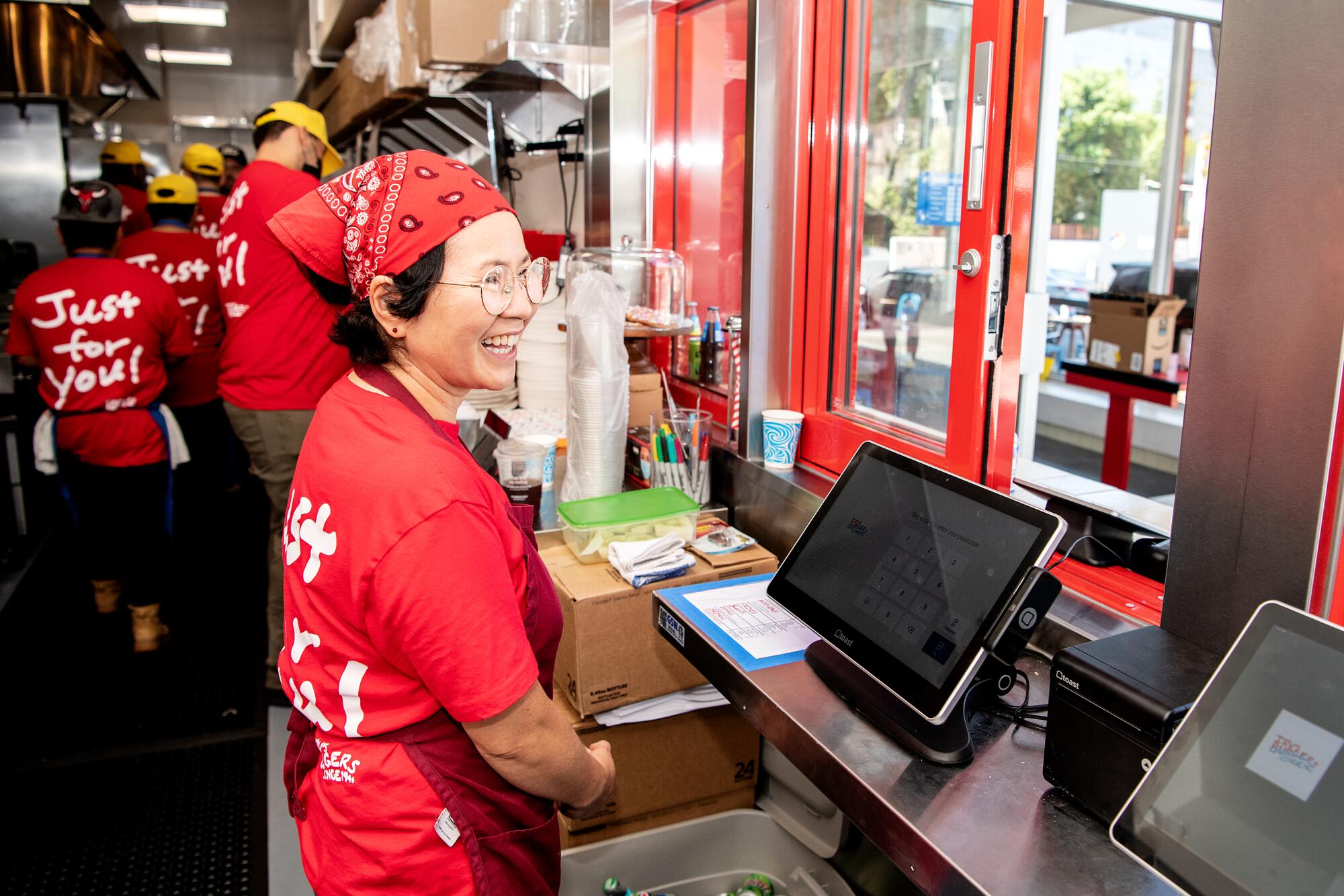 A woman in a red T-shirt and headscarf smiles at guests from behind the register at a hamburger stand.
