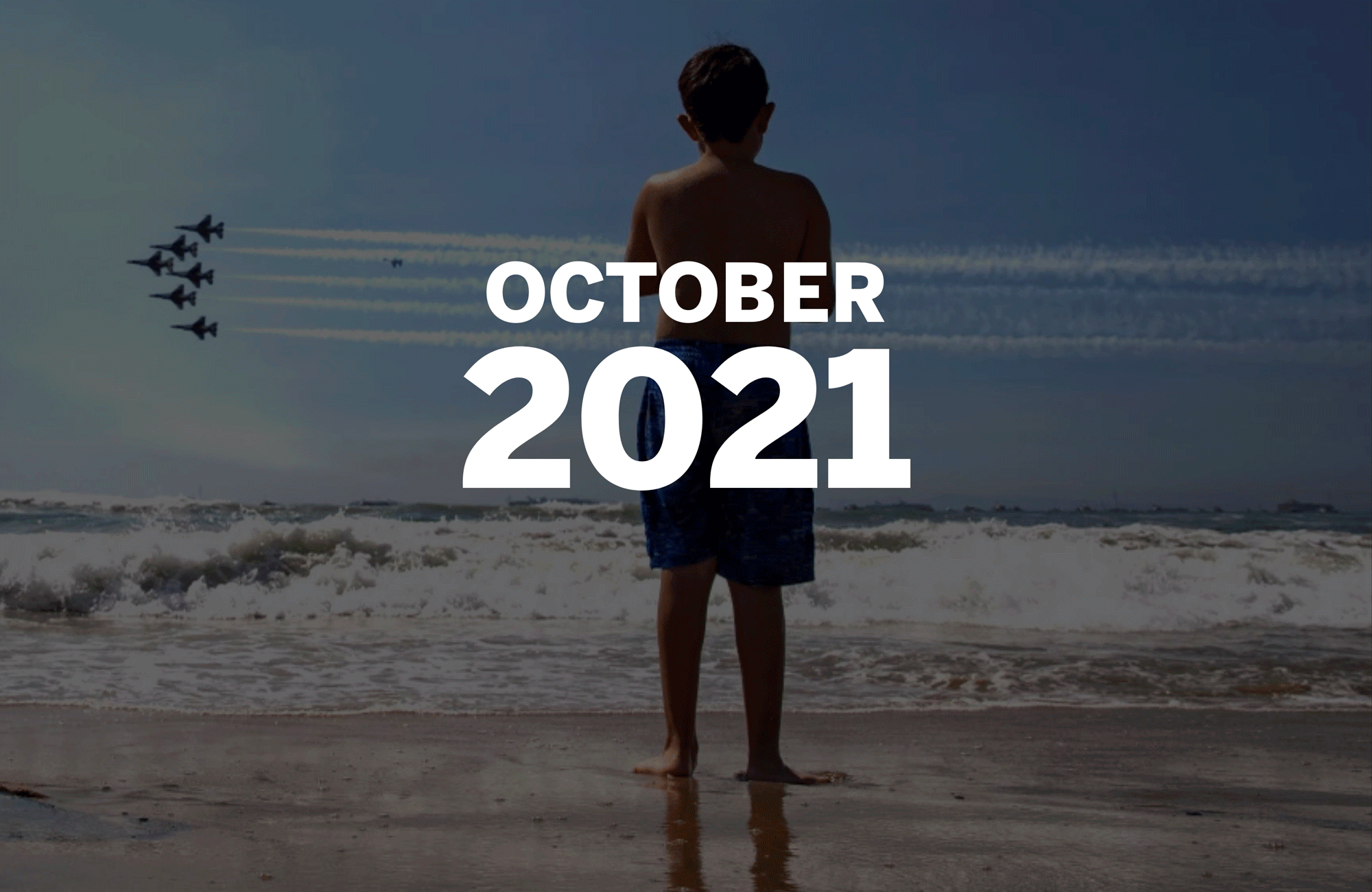 A boy in swim trunks on a beach watches six jets fly in formation