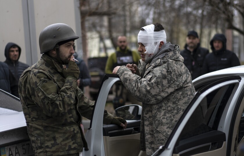 A man in fatigues, his head bandaged, right, stands by his car door as he speaks to another man in fatigues, who is smoking