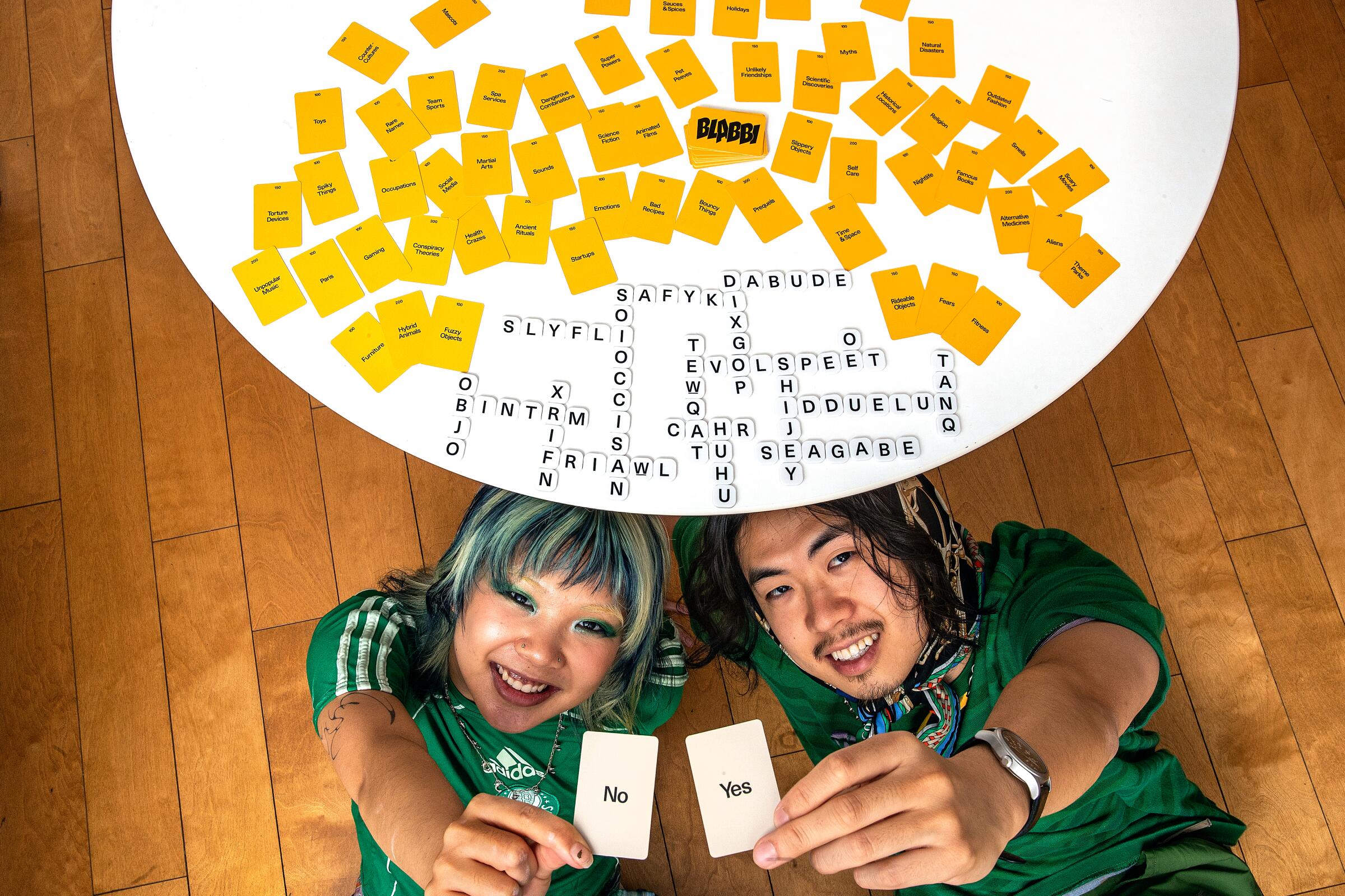 A woman and a man holding up cards that say "no" and "yes" pose under a table where the board game Blabbi is laid out.