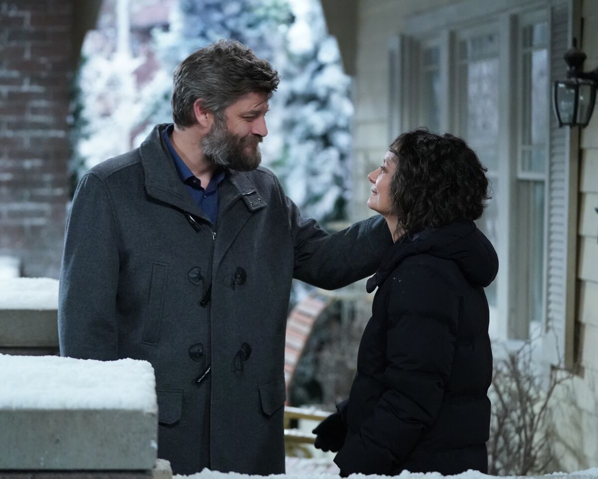 A bearded man and a woman smile at each other in front of a house.