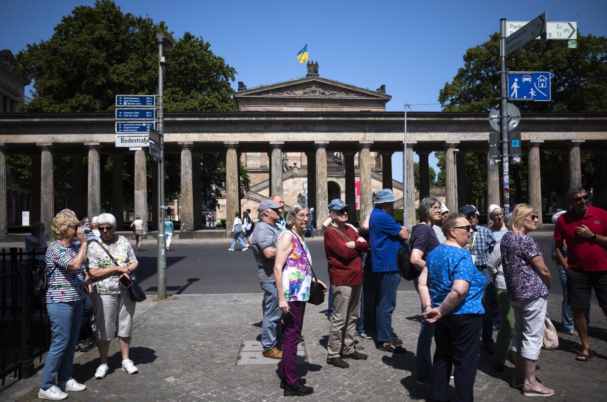 About a dozen tourists standing on a brick walkway near a long line of stone columns, an ornate building beyond them