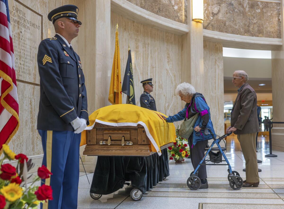 An elderly women rests her hand on the flag draped casket of Bill Richardson as men in uniform flank it.