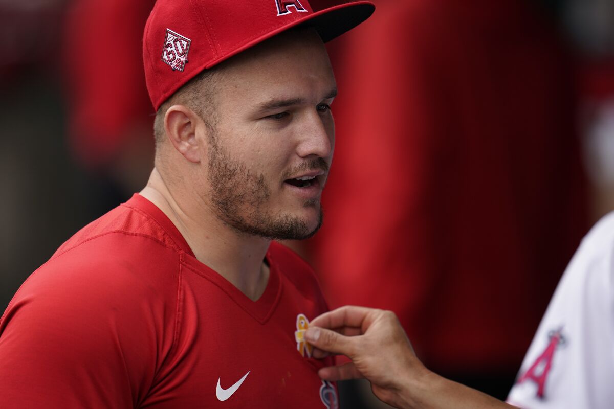 Los Angeles Angels' Mike Trout gets some help to put a yellow ribbon on his shirt in the dugout before a baseball game against the New York Yankees Wednesday, Sep. 1, 2021, in Anaheim, Calif. (AP Photo/Ashley Landis)