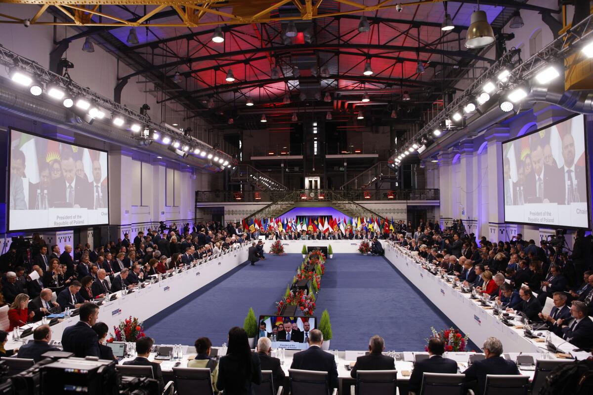People are seated in a rectangular arrangement, flanked by two large TV screens on the walls, with flags at the far end  