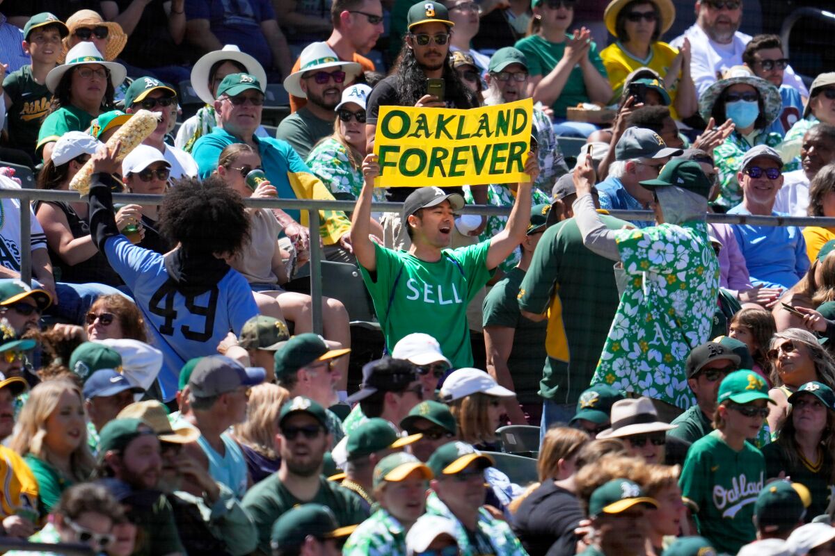 Standing in seated audience a man holds up a sign that reads "Oakland Forever" and wears a T-shirt with a sign "Sell"