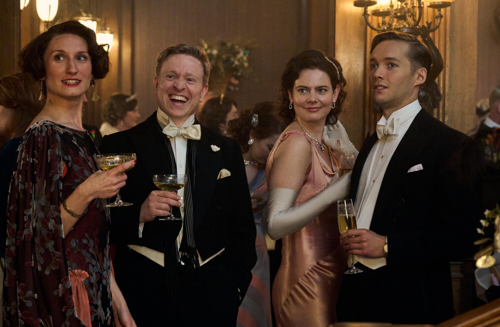 Two couples in formal attire stand holding champagne glasses in a ballroom.