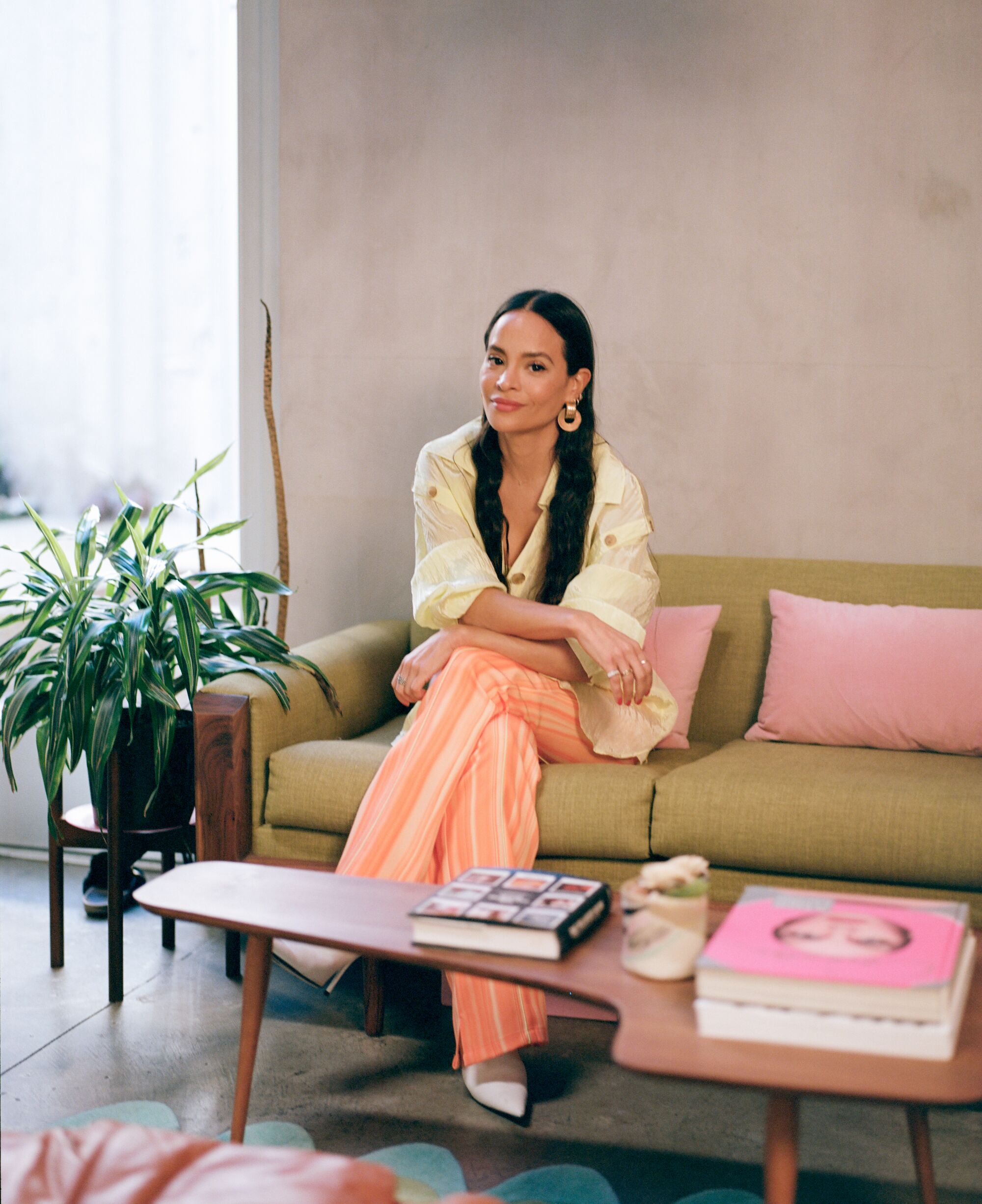 A stylish woman with long dark hair sits on a couch next to a window.