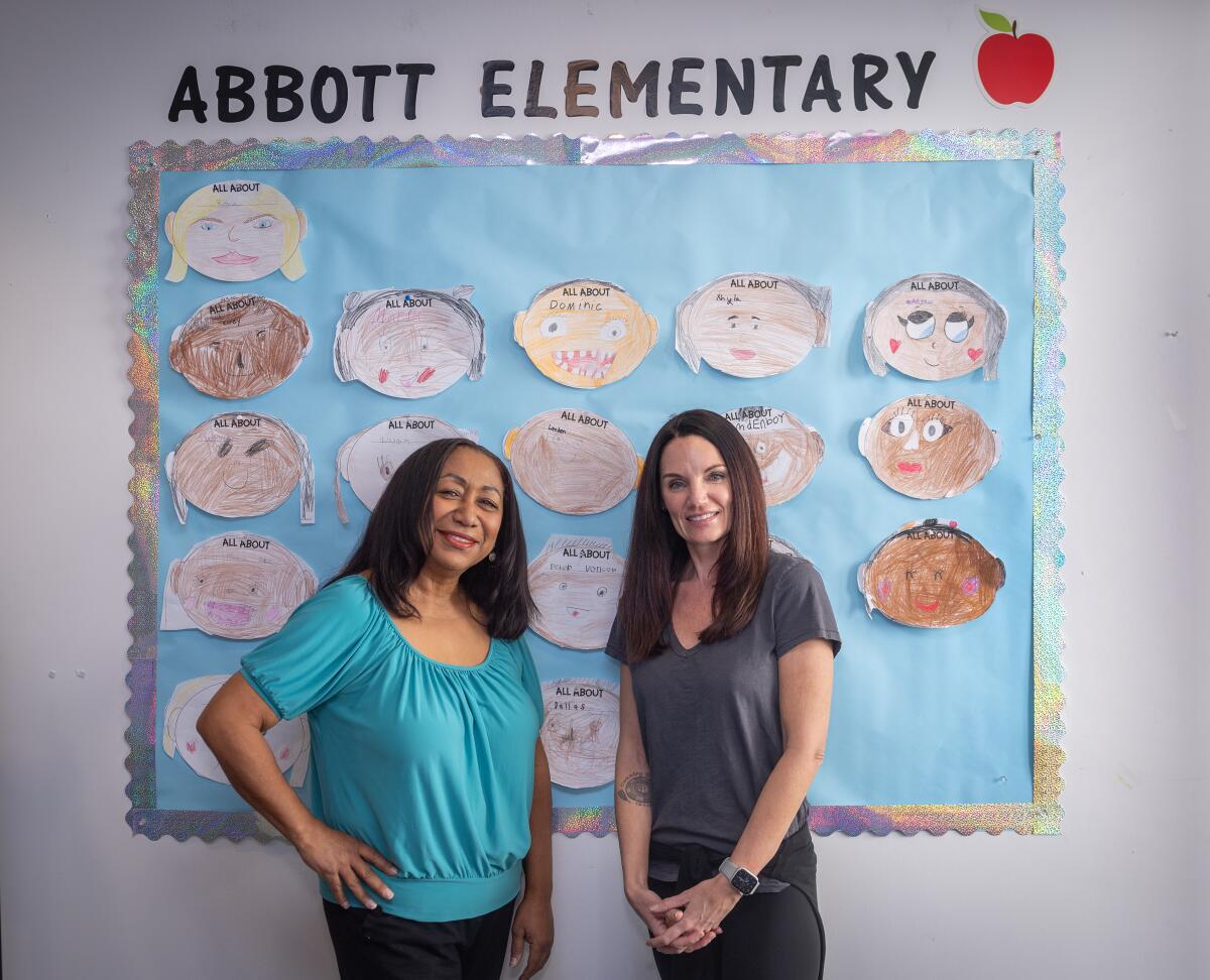 Two women stand and smile in front of a bulletin board decorated with kids' drawings and a sign that says "Abbott Elementary"