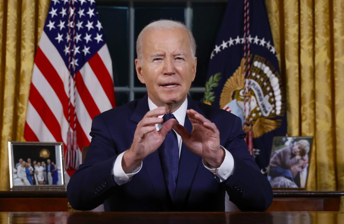 President Biden gestures as he addresses the public from his White House Oval Office desk, flags and family photos behind him