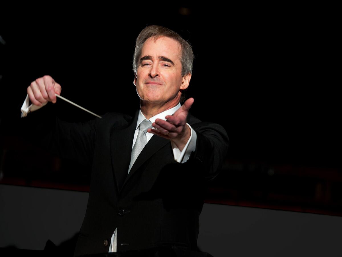 James Conlon, in a black suit and gray tie, holds up a baton in one hand and extends his other hand. 
