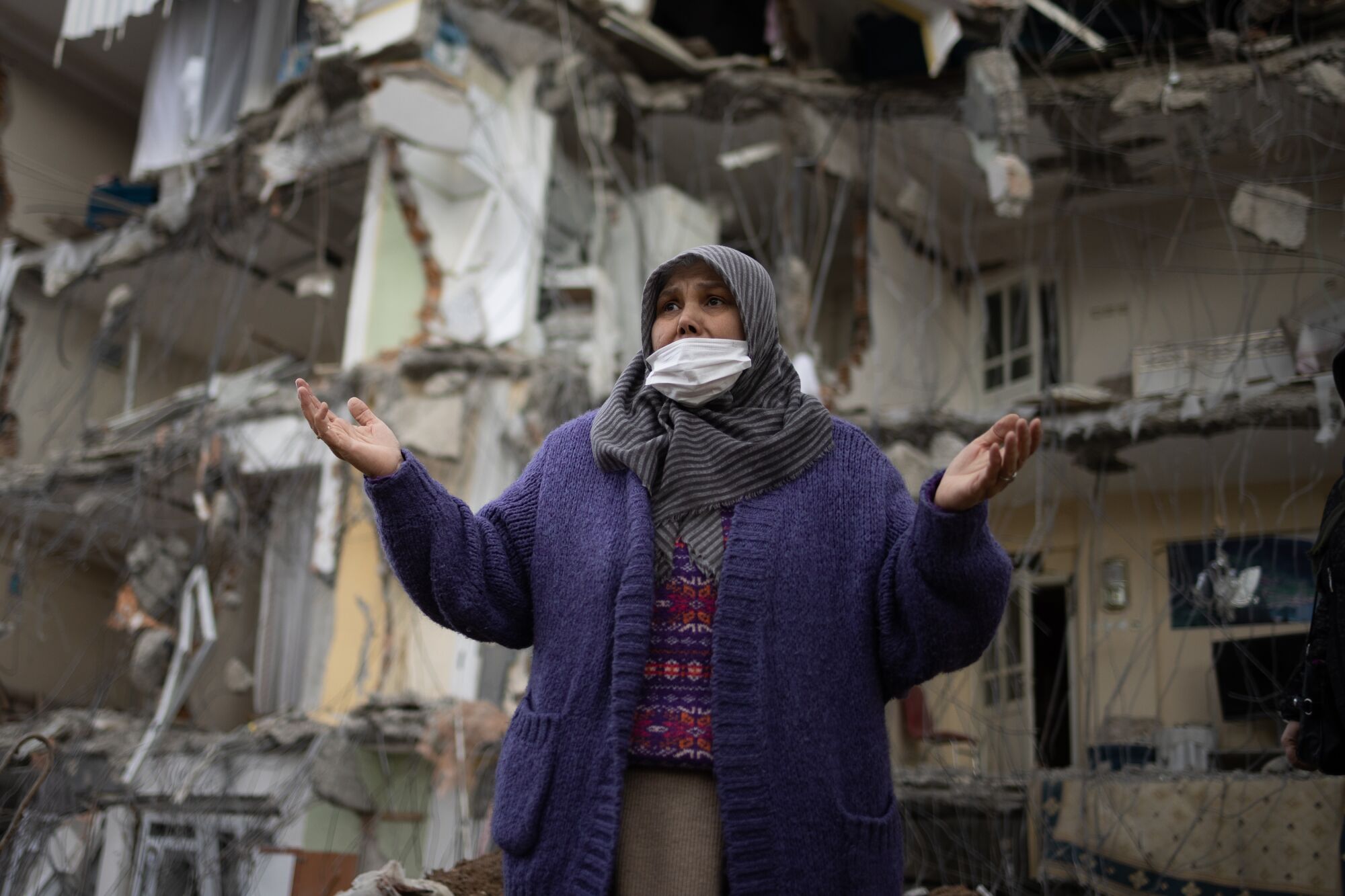 A resident in a head covering holds out her hands in front of her destroyed house.