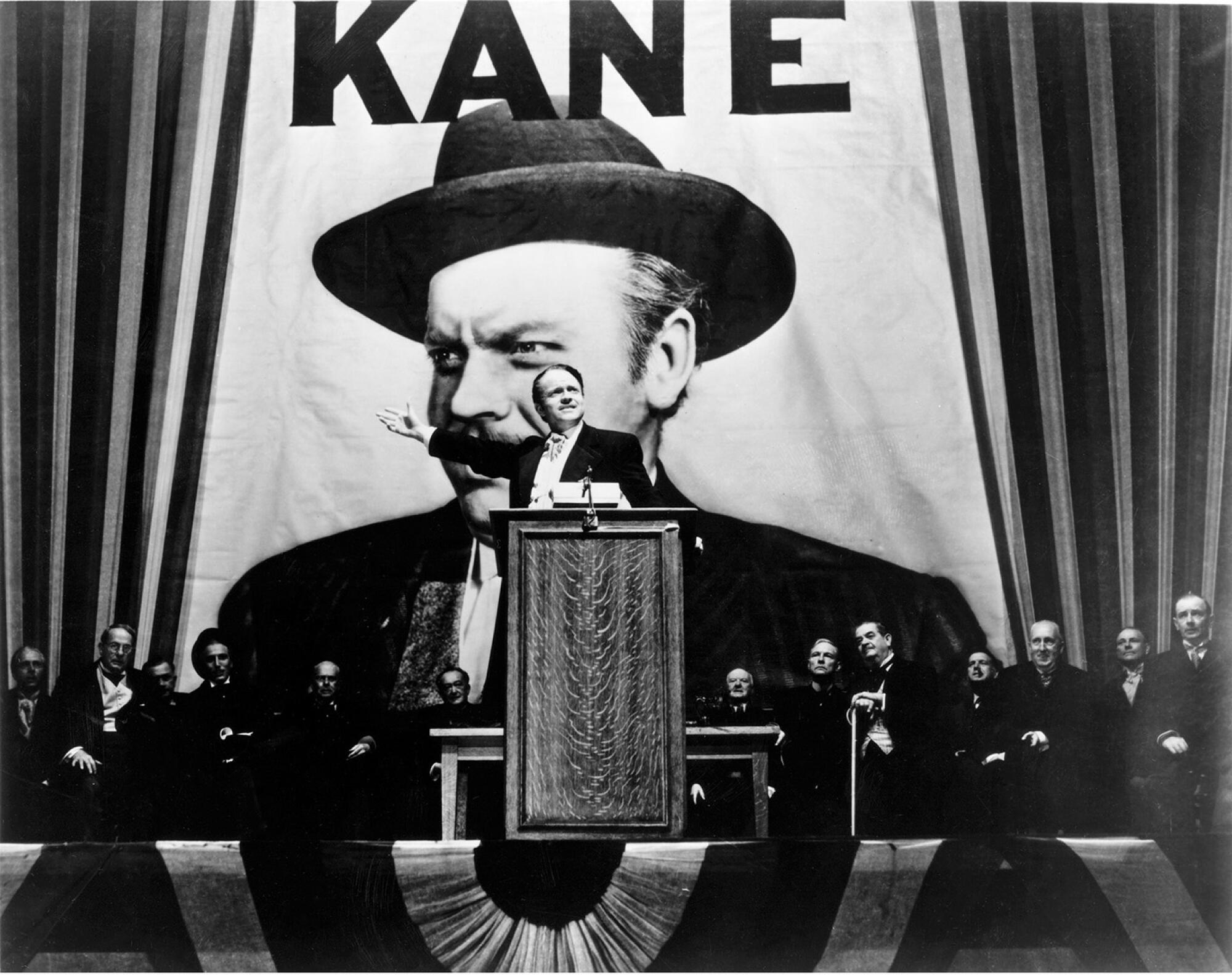 A man delivers a speech from a podium in front of a large picture of himself, marked KANE.