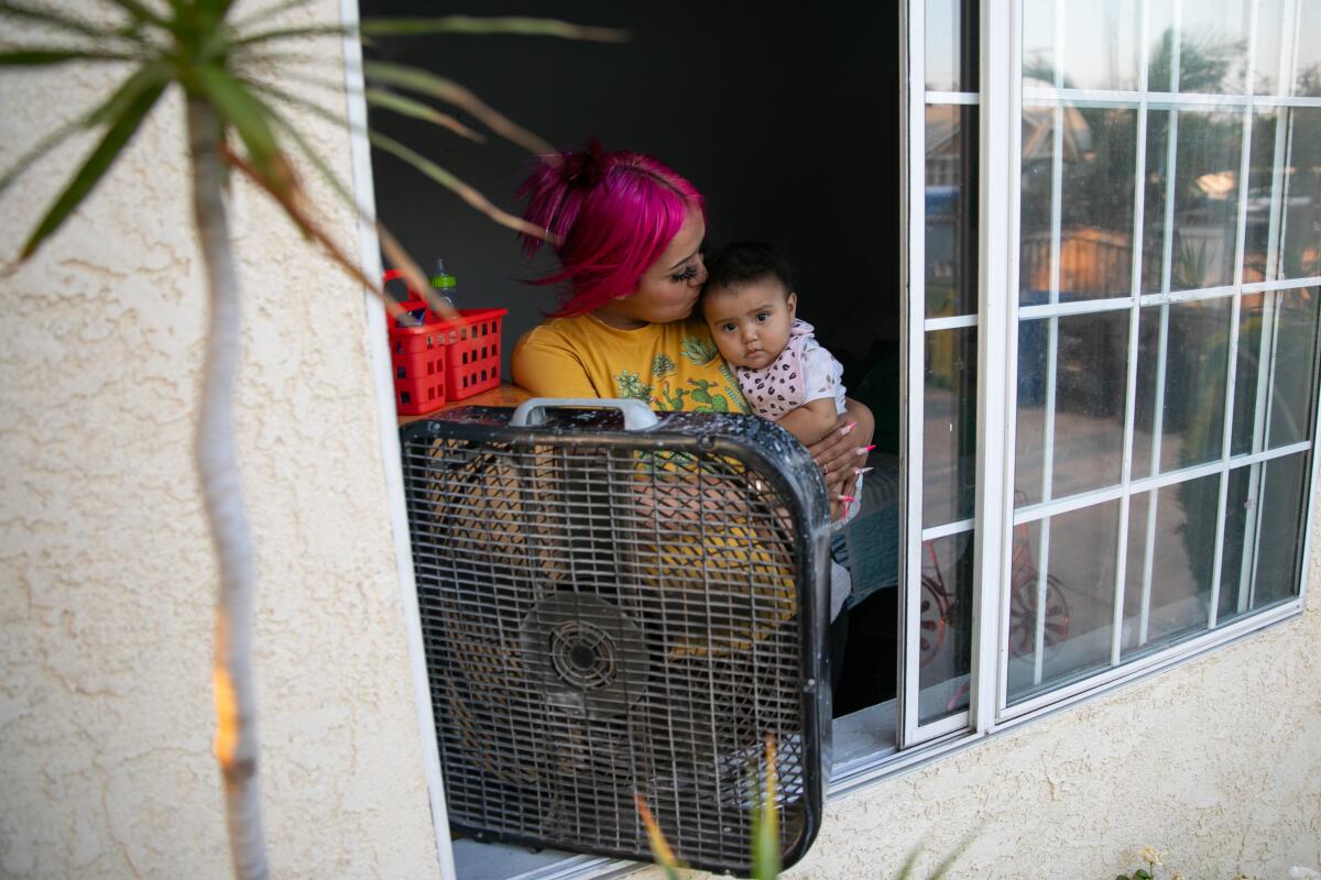 A mother holds her child in front of a fan and an open window.