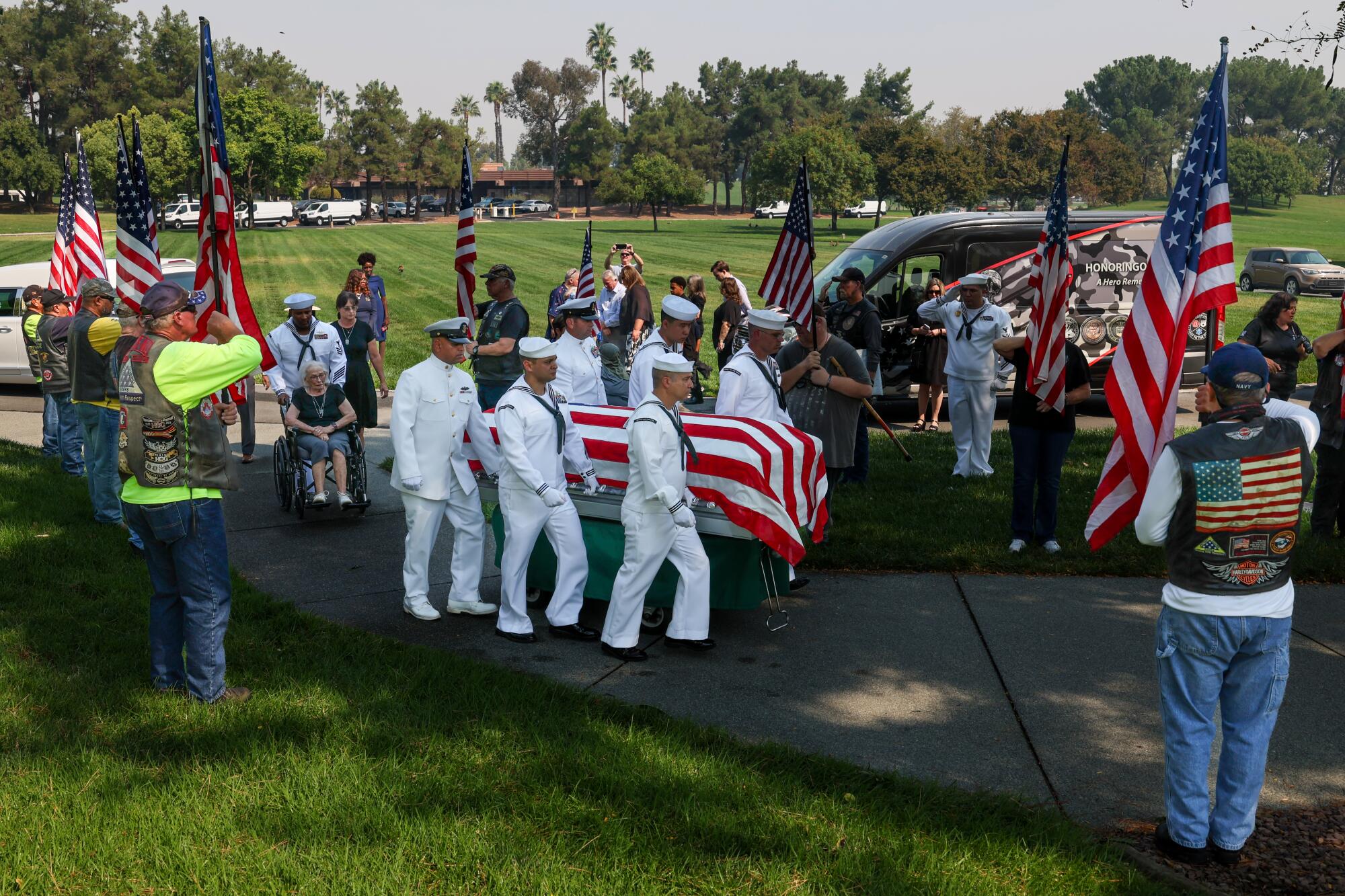 Sailors in white uniform carry a flag-draped casket as others carry U.S. flags around them in a cemetery