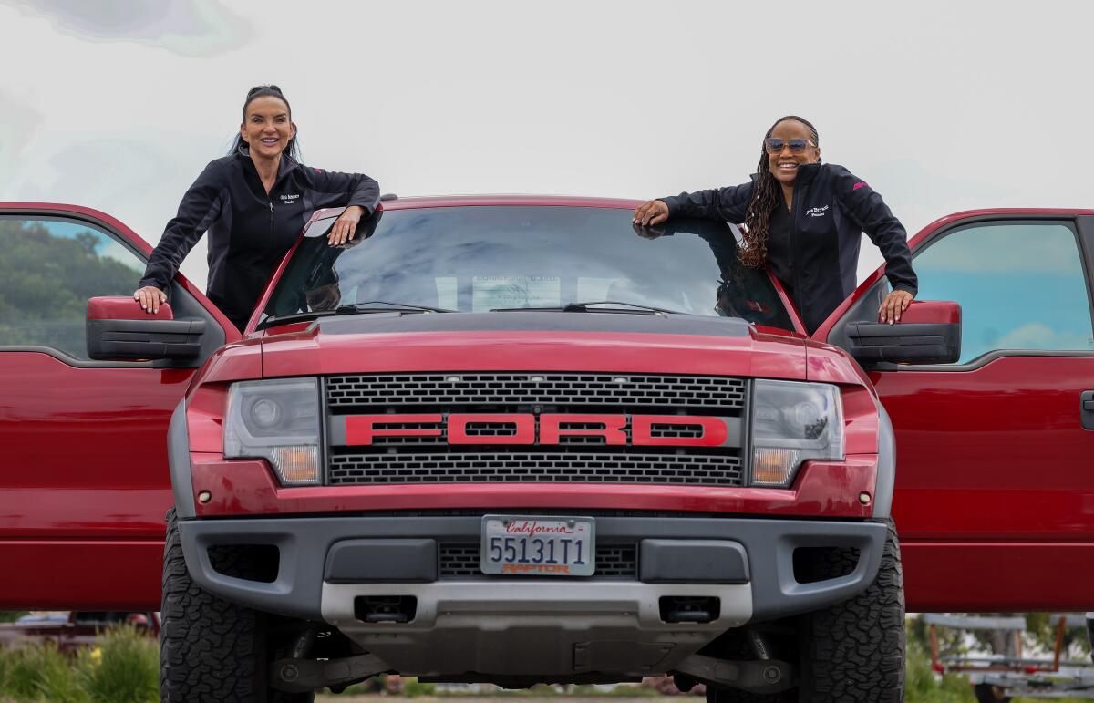 Two women in matching black quarter-zip jackets smile while leaning out the doors of a red Ford truck