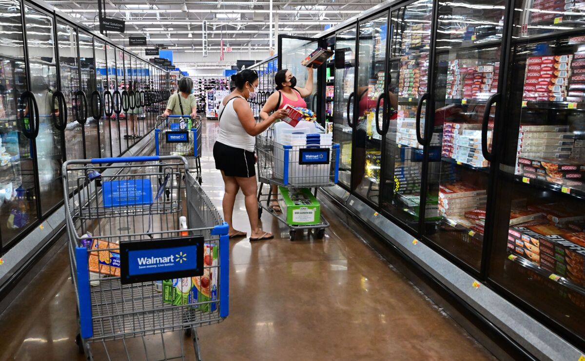 Shoppers with carts stand in front of refrigerated sections for frozen food at a grocery store 