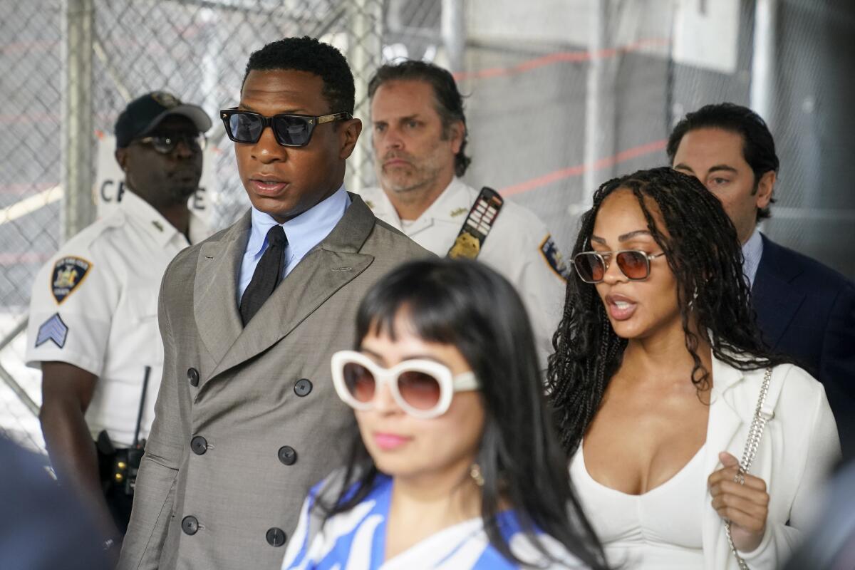 Jonathan Majors in a gray double-breasted suit and sunglasses walking next to Meagan Good in a white dress and jacket