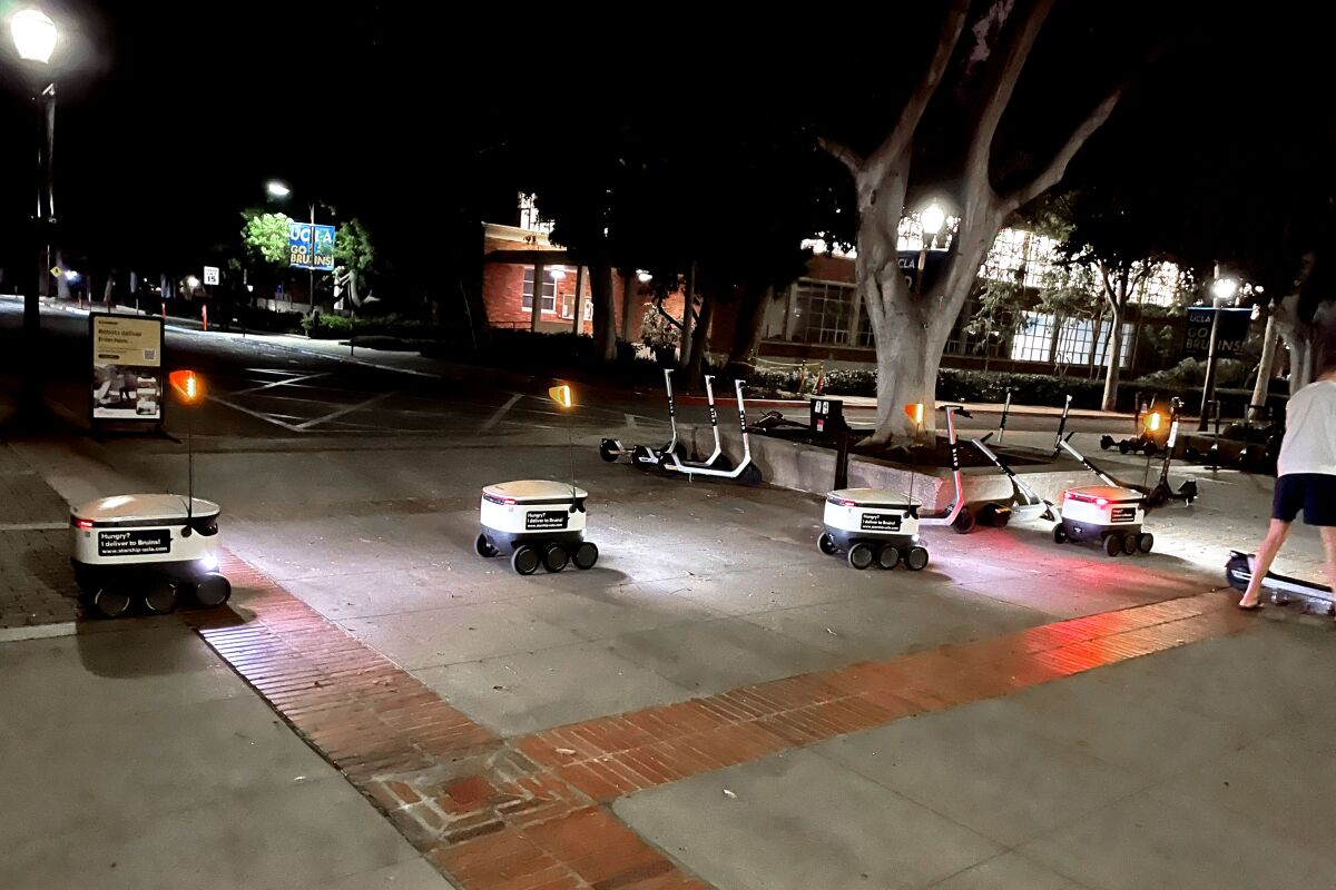 A convoy of food delivery bots rolls across a paved area on UCLA's campus.