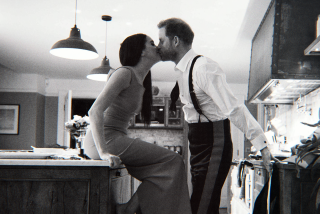 A black-and-white photo of a woman with long dark hair sitting on a counter and kissing a man standing inside a kitchen