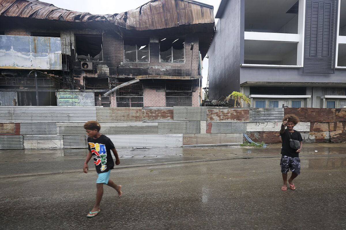 Locals walk past destroyed buildings in Honiara, Solomon Islands, Monday, Dec. 6, 2021. Lawmakers in the Solomon Islands are debating whether they still have confidence in the prime minister, after rioters last month set fire to buildings and looted stores in the capital.(Gary Ramage via AP)