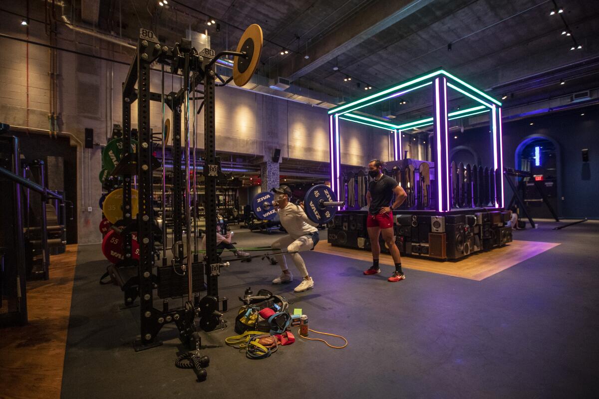 A man does squats with a weighted bar as another man spots him in a gym with a neon-lighted stage in the center