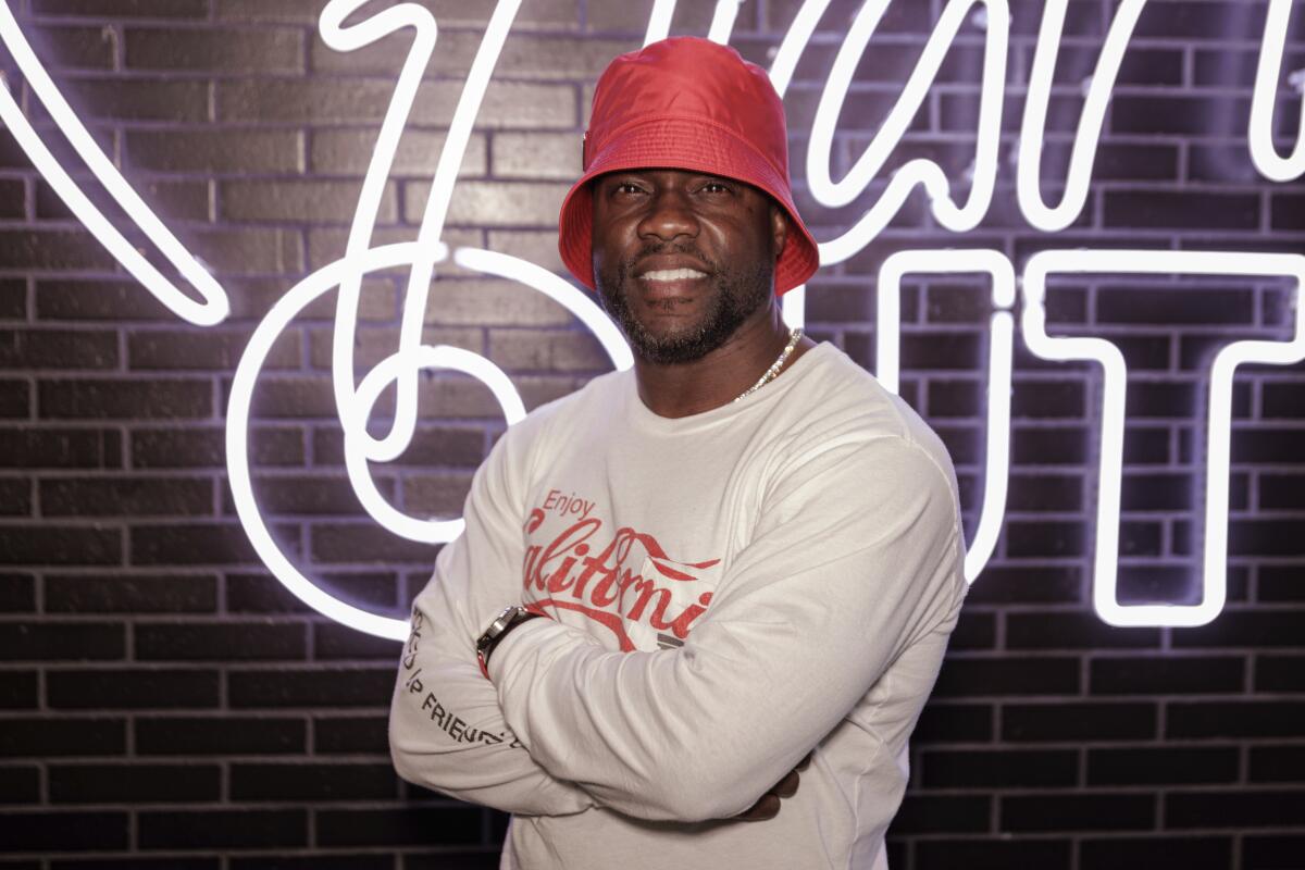 A man in a bucket hat poses in front of a brick wall with a neon sign.