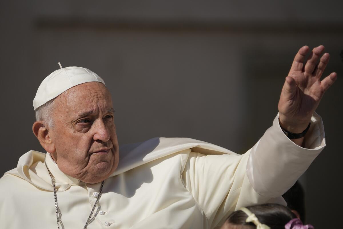 Pope Francis in a white robe and white cap holding up his left hand.
