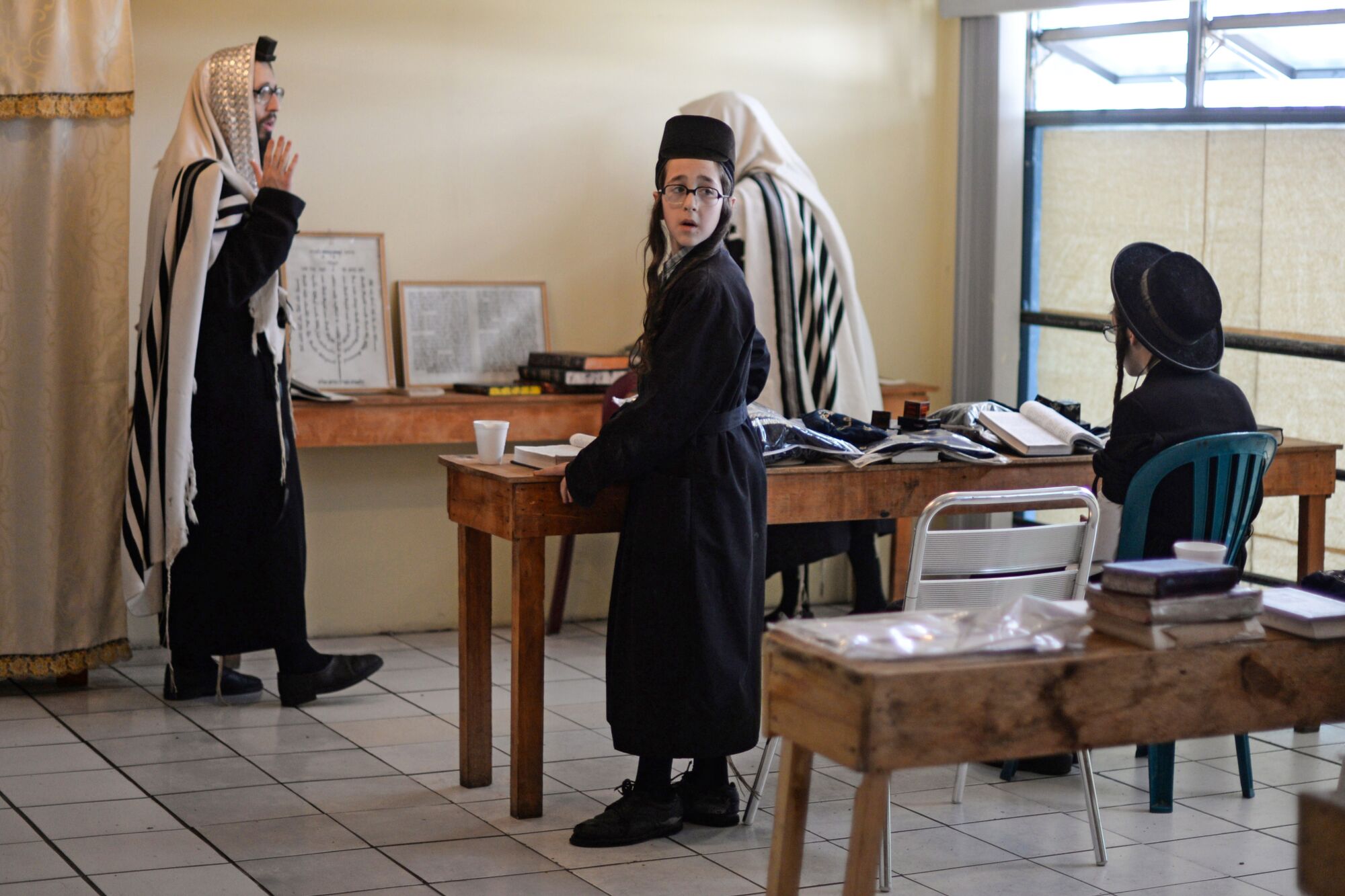 Three people in dark robes stand near two tables. A fourth person, in a dark hat and seated on the right, is reading a book 