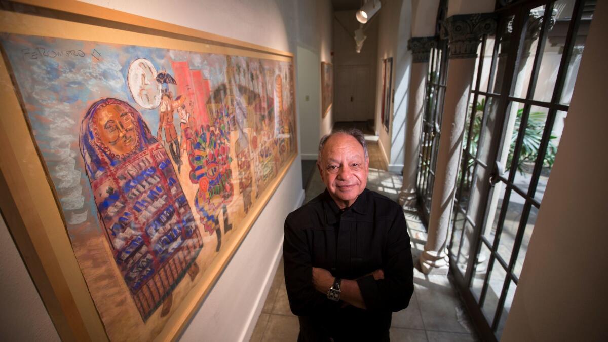 Cheech Marin stands with arms crossed in a hallway next to a large framed painting