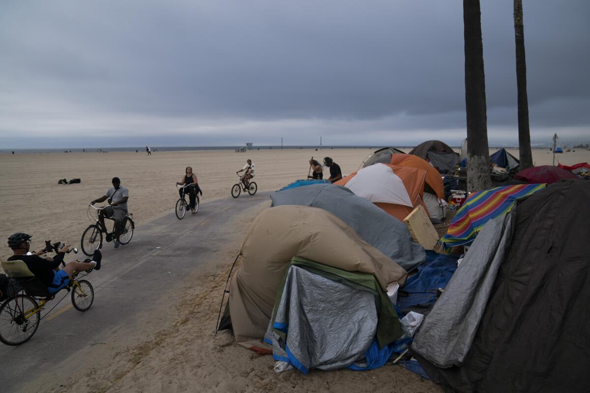 People ride their bikes on a path between the beach and a group of tents.