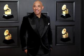 A man in a black tuxedo stands in front of a black wall that has pictures of Grammys on it
