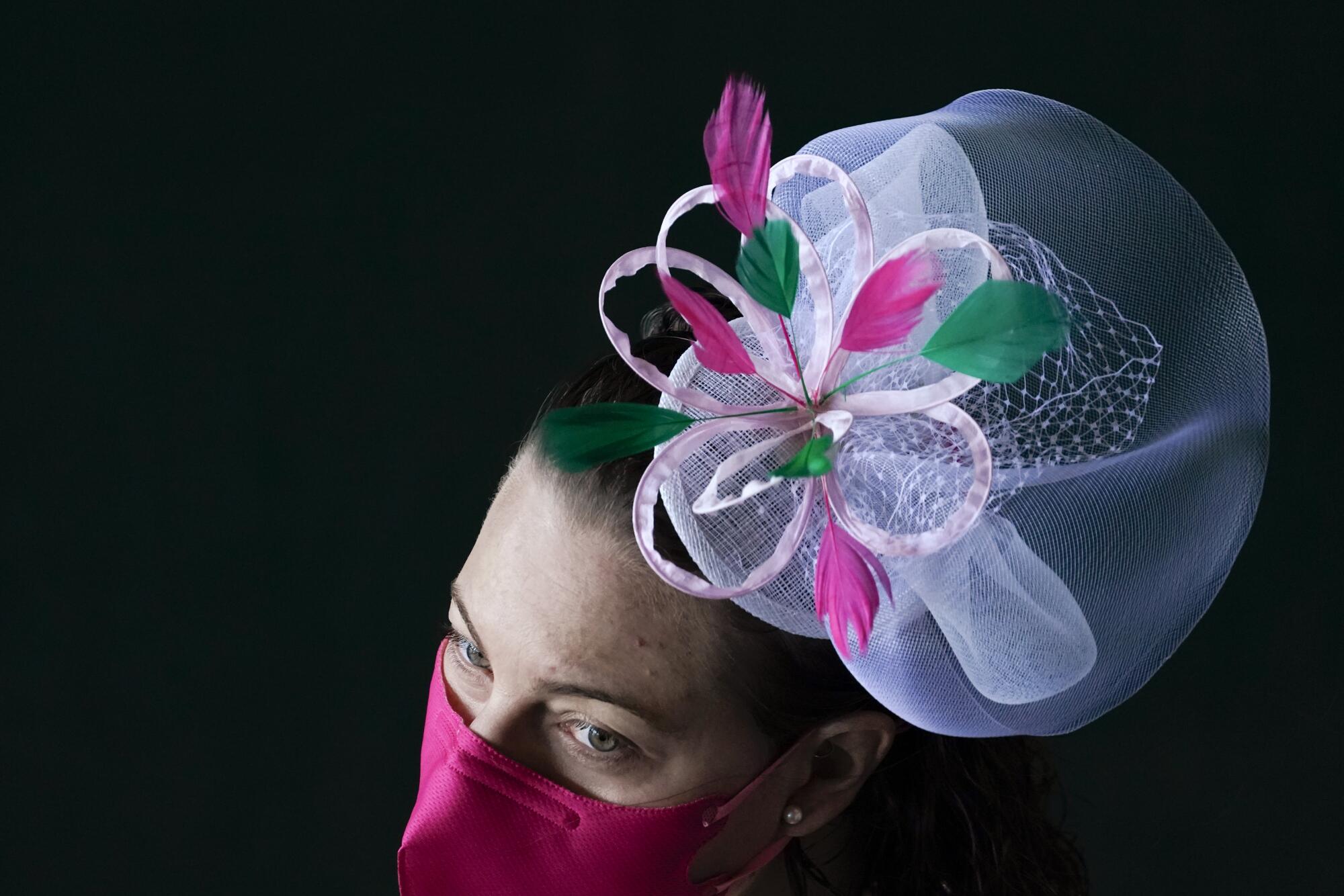 A woman wears a small tulle hat with a pink flower shaped ribbon and green and dark pink feathers, plus a dark pink mask.