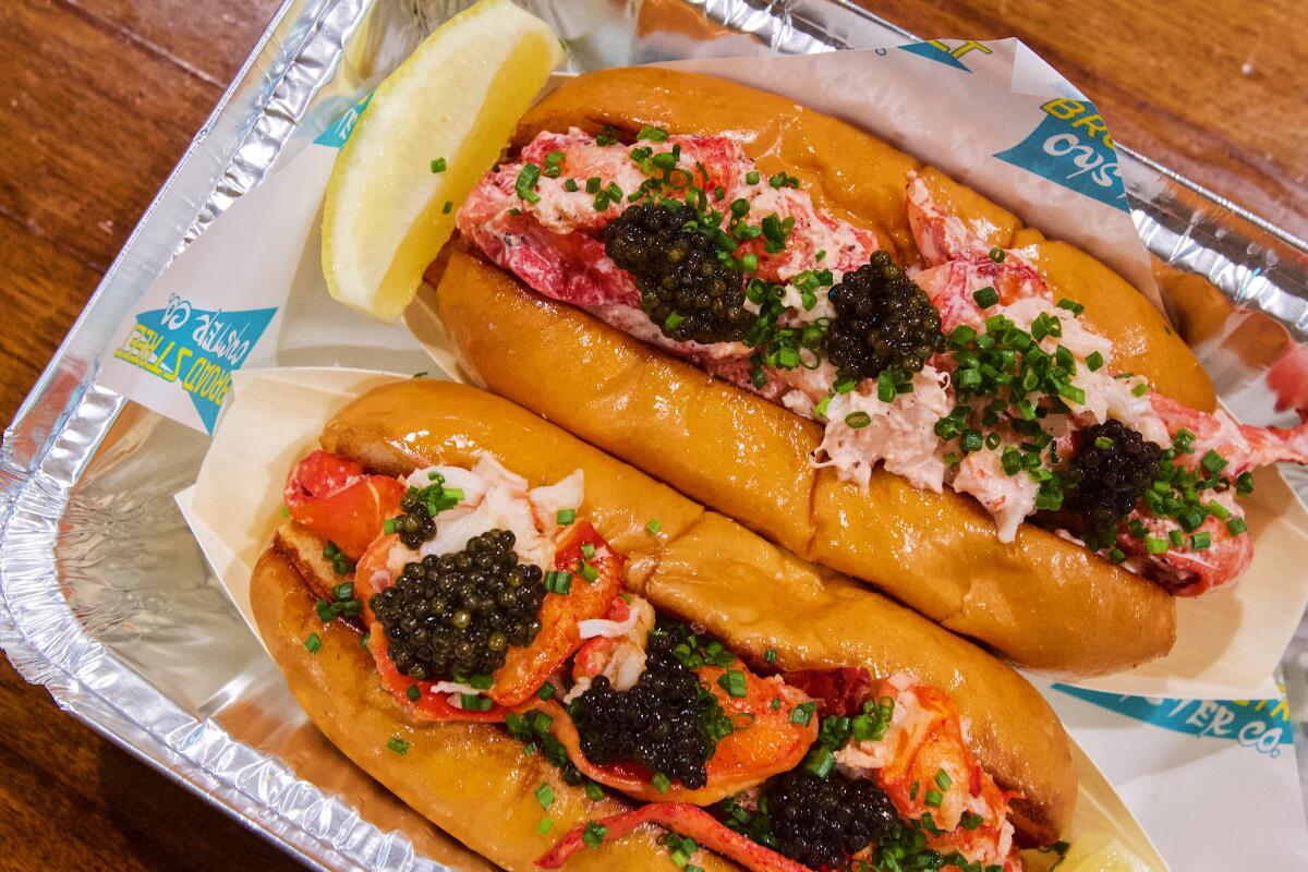 A buttered lobster roll and a chilled lobster roll, both in an aluminum to-go tin and both topped with three bumps of caviar