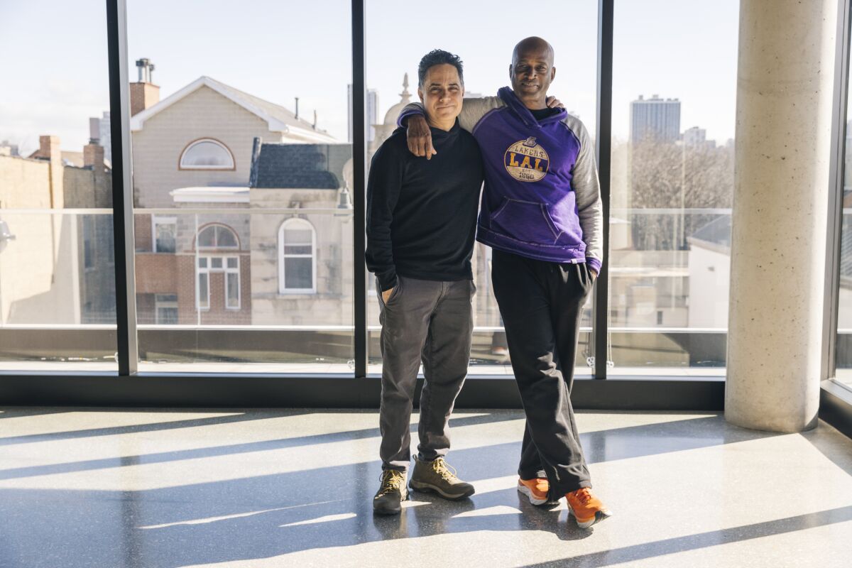 Rajiv Joseph and Kenny Leon stand before a window with views of Chicago with their arms around each other