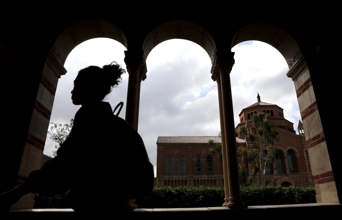 A person is seen in silhouette in a covered walkway with arches, with a view of a red brick building in the background 