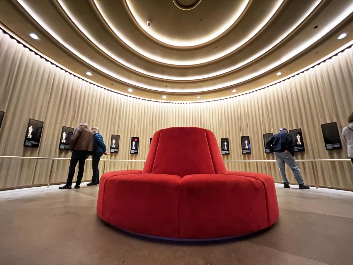 A circular red settee wrapped in a velveteen fabric is seen at the center of a circular gallery with golden walls.