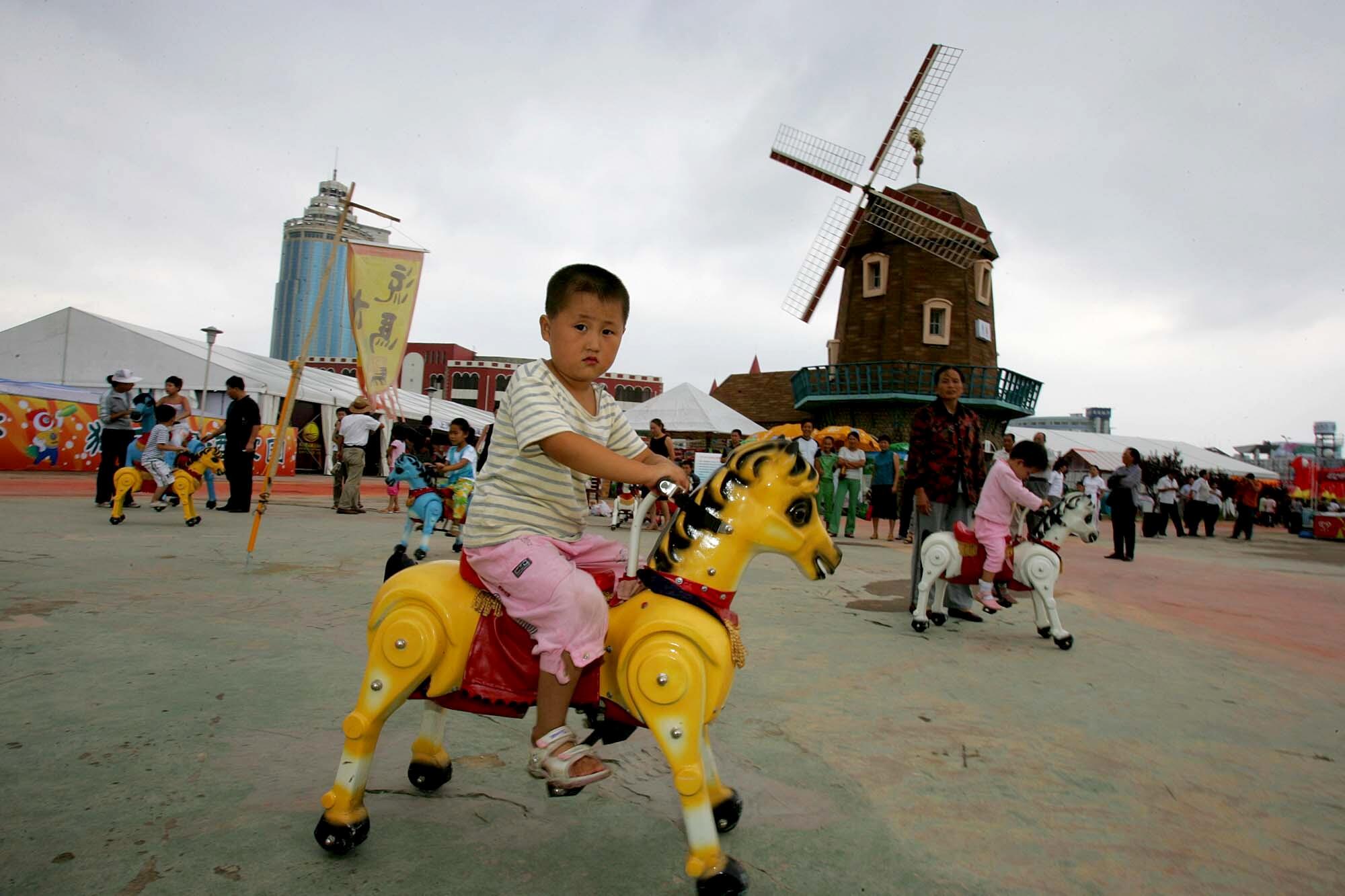 A boy rides a yellow toy pony in a park with a windmill 