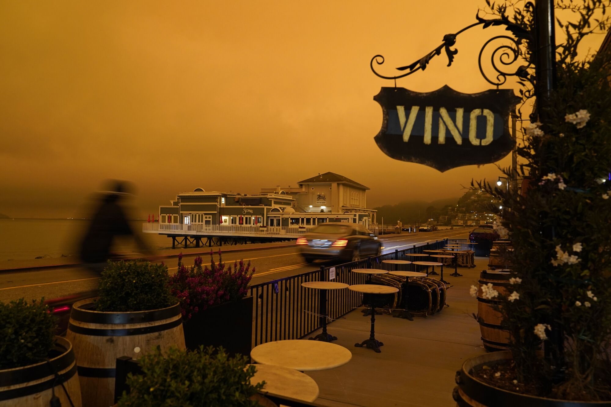 A bicyclist rides past a wine bar's outdoor seating area as smoke darkens the morning sky Wednesday in Sausalito, Calif.