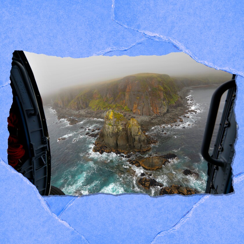 Through helicopter doors, ocean and boulders with green growth are visible.
