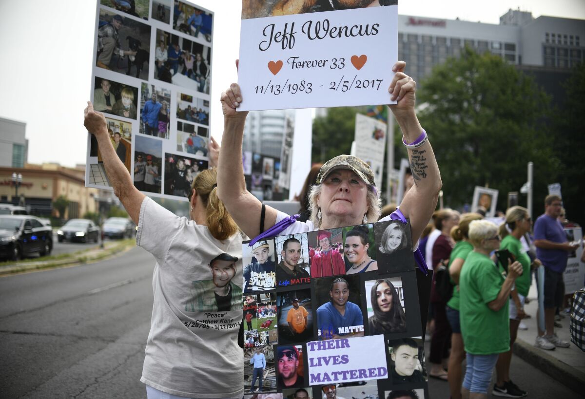 FILE - In this Aug. 17, 2018, file photo, Lynn Wencus of Wrentham, Mass., holds a sign with a picture of her son Jeff and wears a sign of others' loved ones lost to OxyContin and other opioids during a protest at Purdue Pharma LLP headquarters in Stamford, Conn. A landmark settlement in the nation’s opioid epidemic is forcing the owners of OxyContin maker Purdue Pharma to give up the company and pay out $4.5 billion. “Am I happy they don’t have to admit guilt and give up all their money? Of course not,” said Wencus. “But what would that do? It doesn’t bring my son back and it doesn’t help those who are suffering." (AP Photo/Jessica Hill, File)