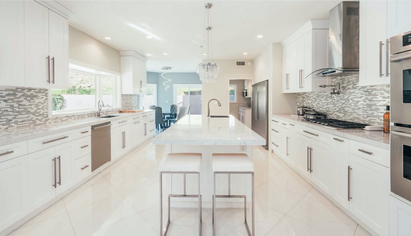 A long kitchen with long, marble-topped center island, white cabinets and tile backsplashes.