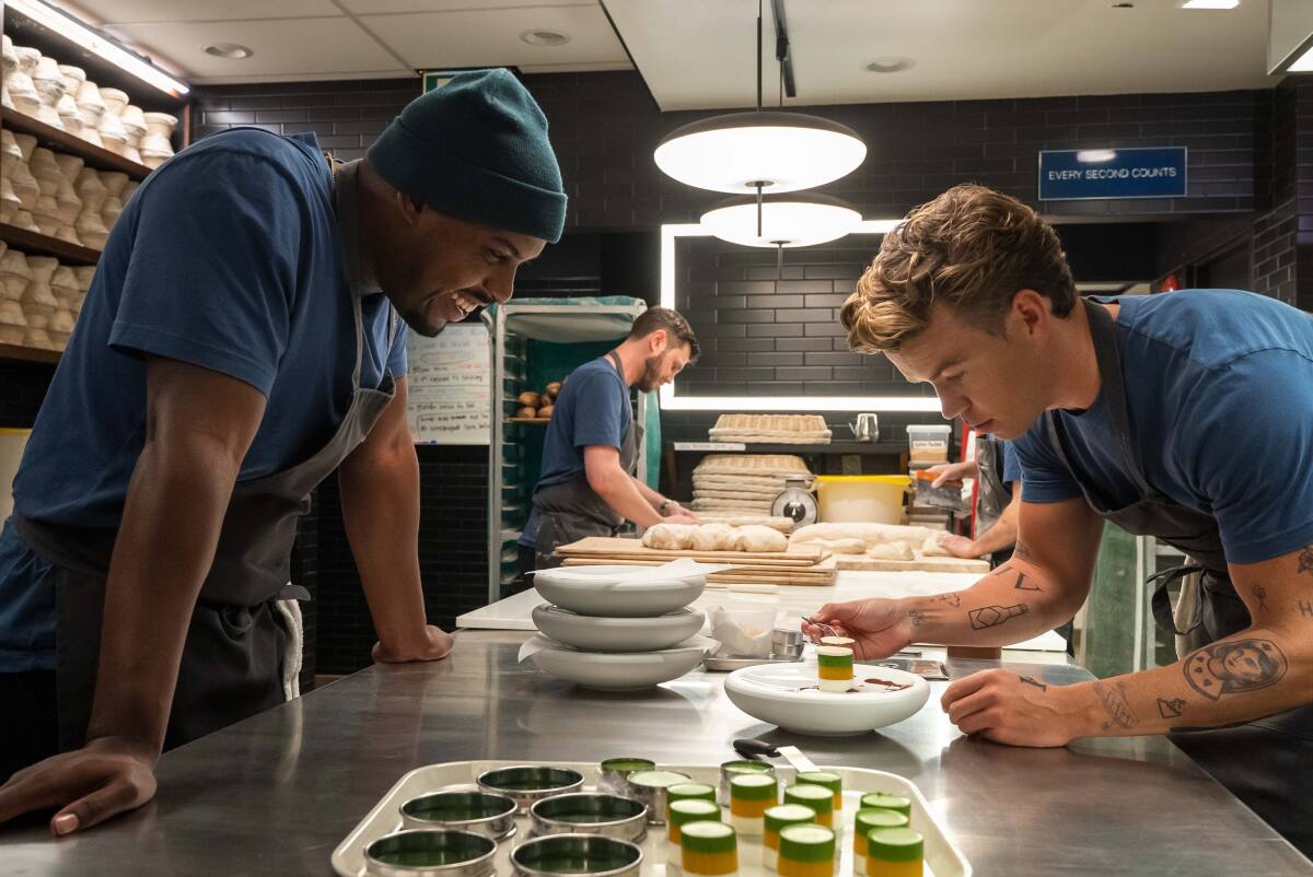 Marcus looks down at a dish that Luca is plating on a stainless-steel kitchen prep table.