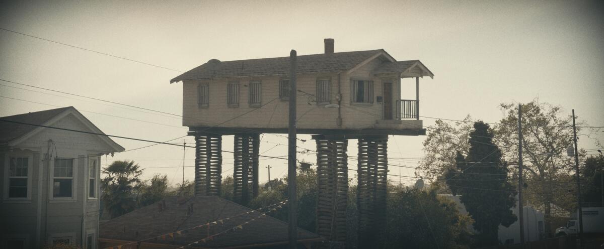 A film still shows a single-story shotgun house elevated on a set of towering stilts.