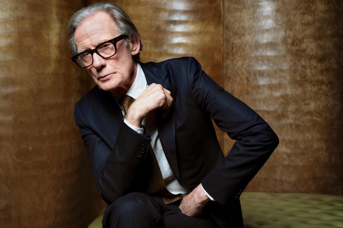 An older man in glasses and a dark suit sits on a bench and leans against a wall for a portrait.